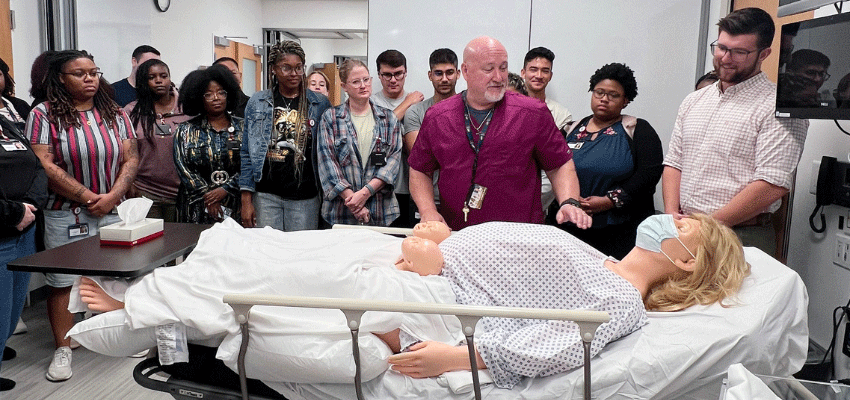 A group of students gathered around a medical mannequin as an instructor demonstrates a procedure in a clinical lab.