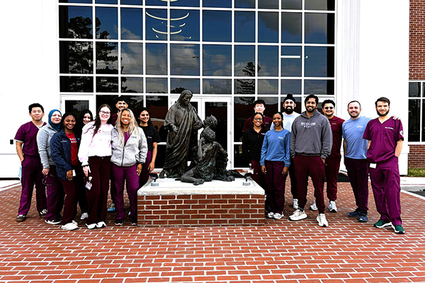 Smiling WCUCOM students in scrubs standing by a statue in front of a campus building.
