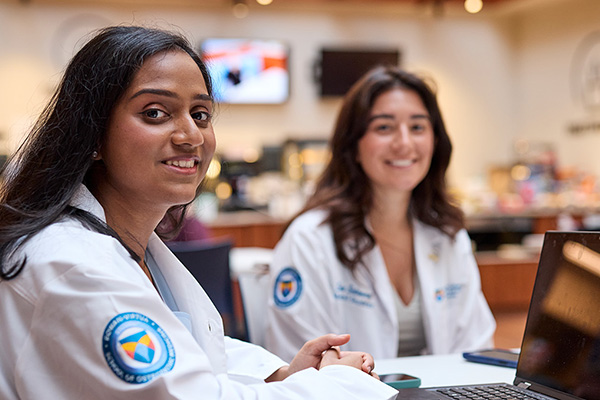 Two female medical students sitting at a table with laptops smile at the camera.