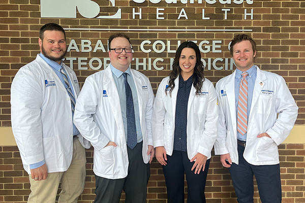 Three smiling male and one female medical students in white coats post in front of a brick wall.