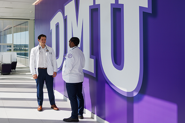 Two male medical students talking in front of a purple wall and large white "DMU" sign.