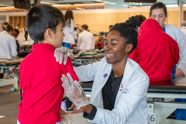 Smiling Black female medical student in white coat checks a boy's arm joint.