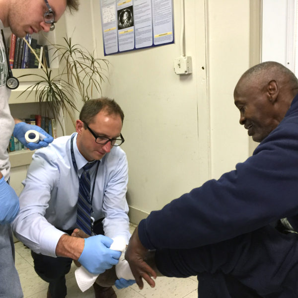 Male Med student observing a professor treat a man's foot wound.