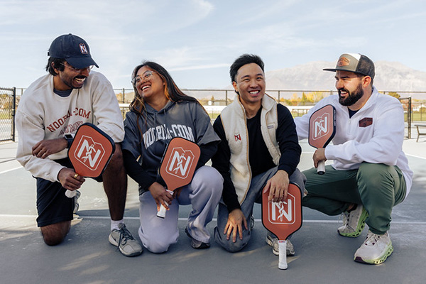 Smiling multiracial group of 4 NoordaCOM students knelling with paddles at a pickleball court