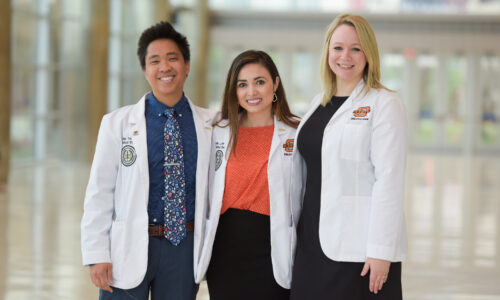 Three diverse OSUCOM students in white coat smiling for the camera