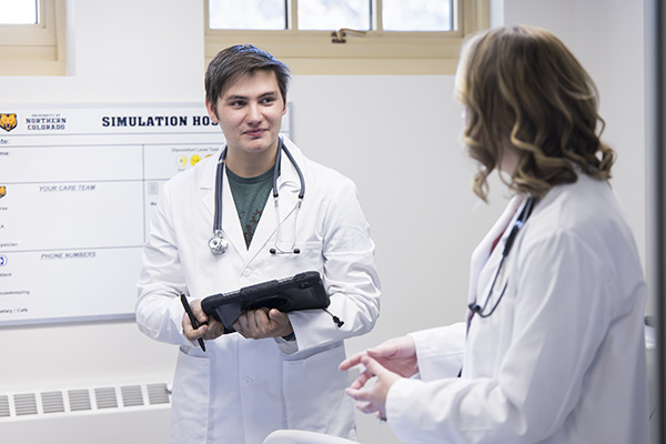 Male medical student in whitecoat, holding a computer and stylus, listens to a female instructor.