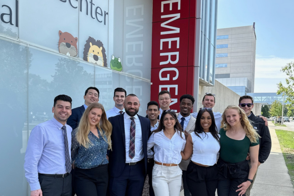 Group of people dressed in business and smart casual attire pose and smile outside a medical center near an “emergency” sign on a sunny day.