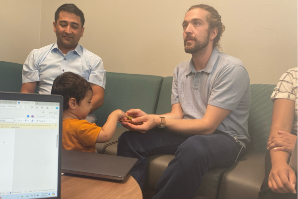 A child in an orange shirt interacts with a bearded man holding a small toy on a couch. A laptop is visible on a table, and two adults sit nearby, one watching the child warmly.