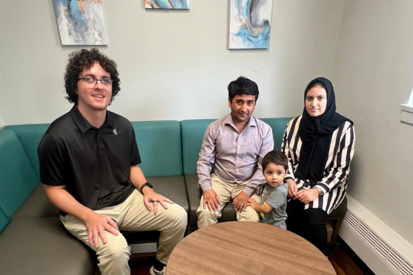 A group of four people, including a young child, sit on a teal corner sofa. The room has light walls with abstract art. The atmosphere is calm and relaxed.