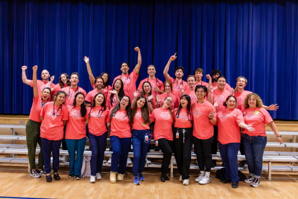 A group of diverse individuals wearing matching pink shirts poses enthusiastically on bleachers. The mood is celebratory against a blue curtain backdrop.