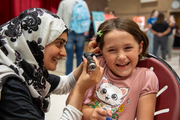 A woman wearing a patterned hijab uses an otoscope to check a smiling girl's ear in a playful setting. The girl wears a pink cat t-shirt.