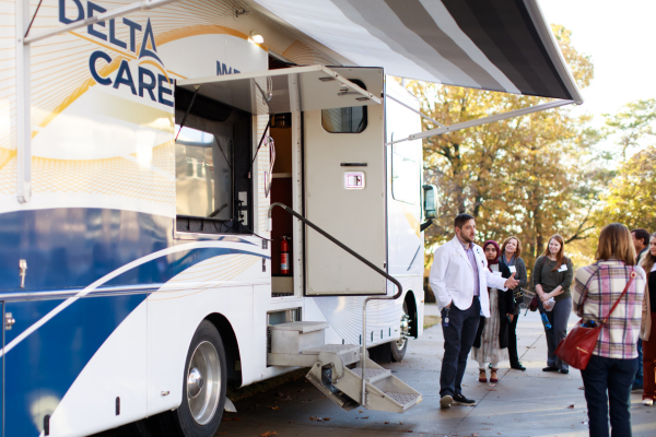 A healthcare professional speaks to a group outside a mobile clinic labeled 