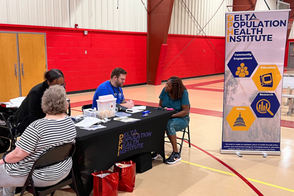 A community health event indoors with a table for the Delta Population Health Institute. Four people engage in discussion, with a banner promoting community engagement, education, research and policy.