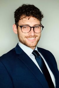 A man in a suit and glasses smiles in a professional headshot against a neutral background.