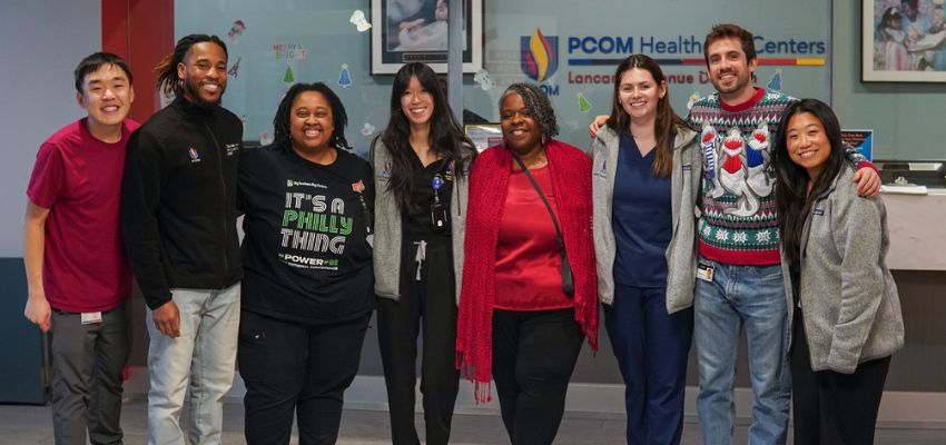 A diverse group of healthcare workers and students stand smiling together inside a PCOM Health Center.