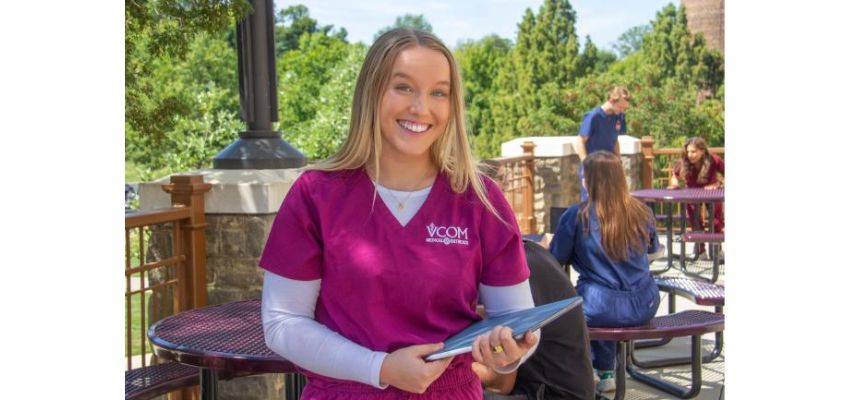 A medical student in maroon scrubs smiles while holding a tablet on an outdoor campus patio with other students in the background.