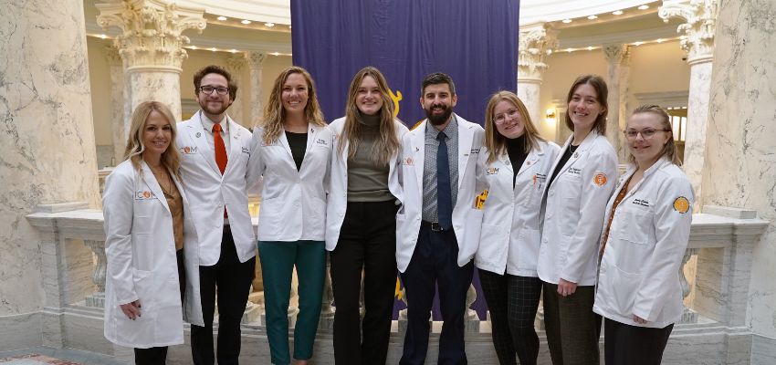 A group of eight medical students in white coats pose inside a government building with tall marble columns.