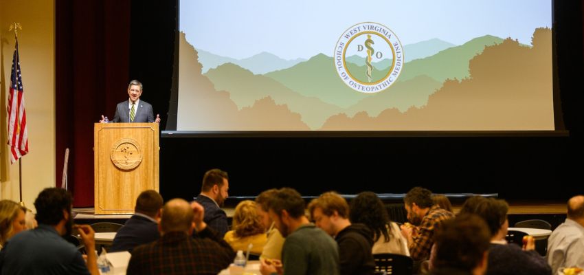 A speaker addresses a seated audience at a podium with the WVSOM logo, in front of a large screen displaying a scenic mountain graphic.