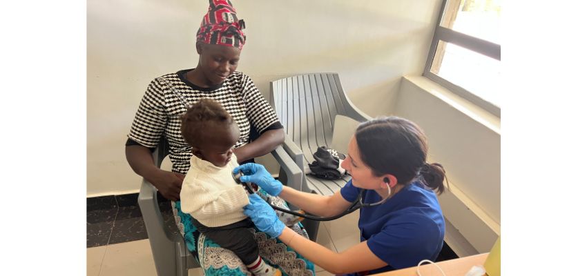 A student in blue scrubs uses a stethoscope to examine a baby sitting on their mother’s lap in a clinic setting.