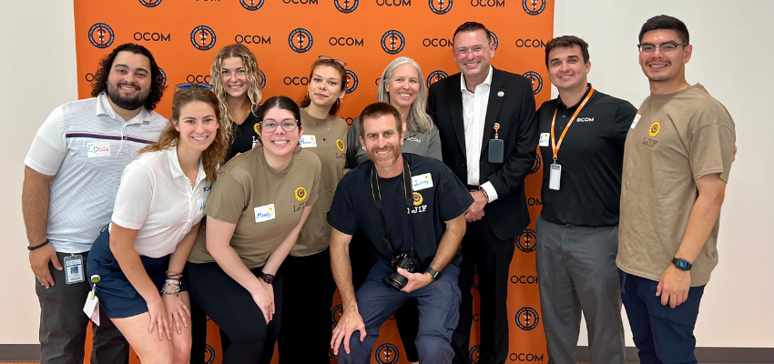 A group of students and faculty pose together in front of an OCOM step-and-repeat banner, smiling during a campus or event gathering.