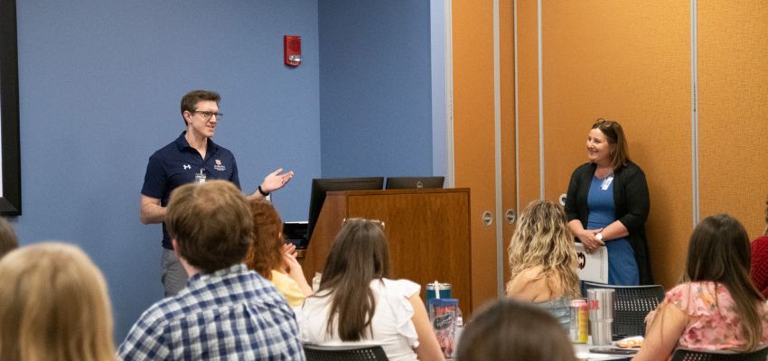 A man presents to a room of seated attendees during a classroom discussion or meeting.