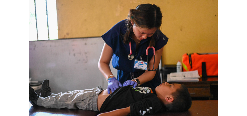 A medical student examines a young boy's abdomen during a clinical outreach or service trip in a rural setting.