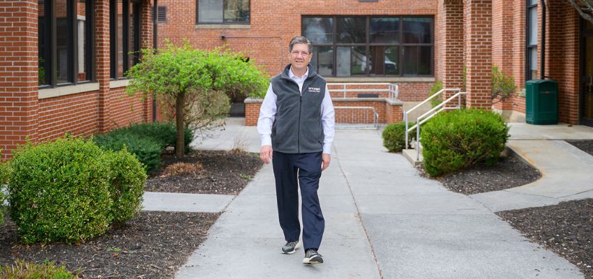 A man walks confidently along a pathway on a campus with red-brick buildings and green landscaping.