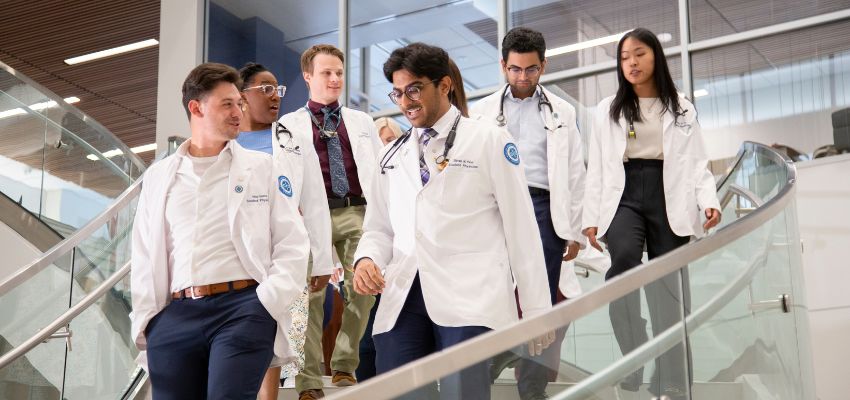Seven medical students in white coats walk down a staircase inside a modern academic building.