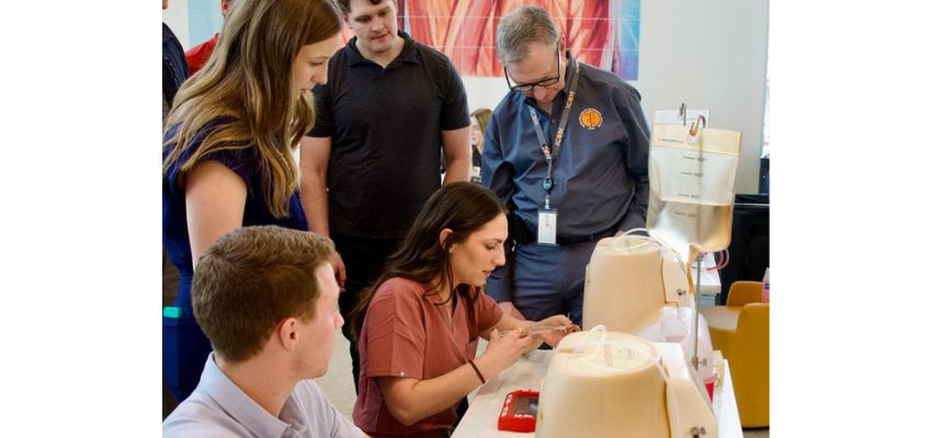 A medical student practices a hands-on technique using simulation equipment as peers and a faculty member observe during a clinical skills training session.