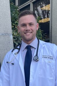PCOM student doctor Patrick Fitzgerald smiles outdoors in his white coat with a stethoscope draped around his neck.