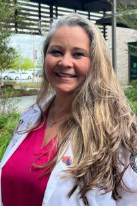A woman wearing a white coat and pink top stands smiling outdoors near a building and greenery.