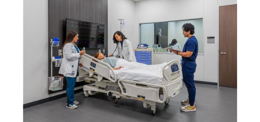 Three UNTHSC-TCOM students and a faculty member stand around a hospital bed, engaging in a simulated patient care exercise in a modern clinical skills lab.