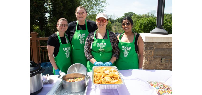 Four people wearing matching green aprons stand behind a table with food trays and serving utensils outdoors.
