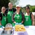 Fourpeoplewearingmatchinggreenapronsstandbehindatablewithfoodtraysandservingutensilsoutdoors