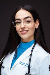 Shahrzad smiles for a professional photo, wearing her whitecoat and stethoscope.