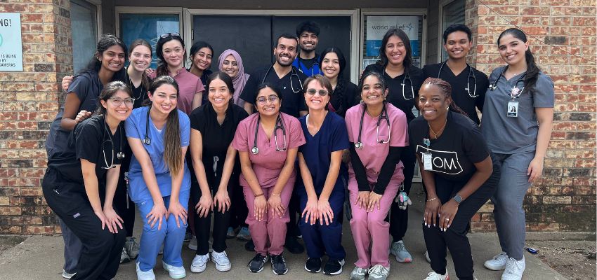 A group of UNTHSC-TCOM medical students gather in front of a clinic building, smiling in their scrubs with stethoscopes.