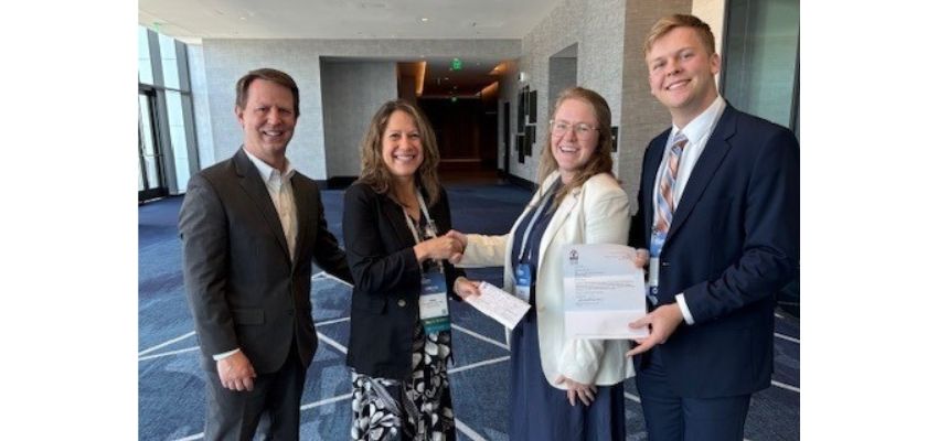 Four individuals in formal attire, including VCOM-Carolinas representatives, pose together at a conference while holding a certificate.