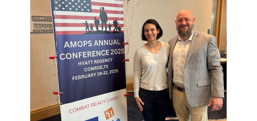 Two VCOM-Louisiana attendees stand beside a banner promoting the AMOPS Annual Conference 2025 in Conroe, Texas.