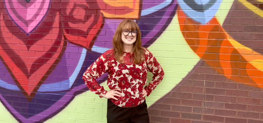 A young woman in a floral blouse stands smiling in front of a colorful mural painted on a brick wall.