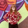 Ayoungwomaninafloralblousestandssmilinginfrontofacolorfulmuralpaintedonabrickwall