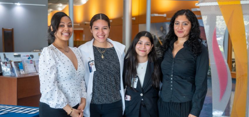 Four female students dressed in business or medical attire smile for a photo in a bright lobby area.