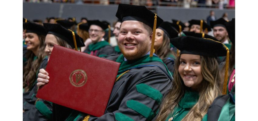 A male graduate in regalia holds up his diploma and smiles during a commencement ceremony.