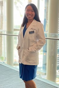 A medical student in a white coat smiles while posing in a brightly lit indoor space.