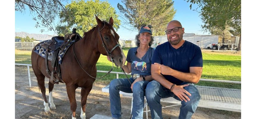 Two people sit on a bench next to a saddled horse, smiling in a sunny outdoor setting.