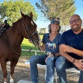 Twopeoplesitonabenchnexttoasaddledhorsesmilinginasunnyoutdoorsetting