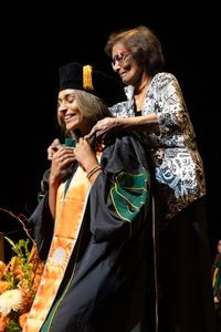 A medical graduate is being hooded during a graduation ceremony, smiling as a family member assists her on stage.