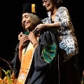 Amedicalgraduateisbeinghoodedduringagraduationceremonysmilingasafamilymemberassistsheronstage