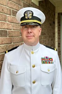 A man in a U.S. Navy dress white uniform stands in front of a brick wall, looking directly at the camera.