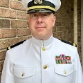 AmaninaUSNavydresswhiteuniformstandsinfrontofabrickwalllookingdirectlyatthecamera