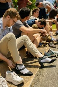 A group of students sits on a curb putting on women's heels, participating in an awareness event.